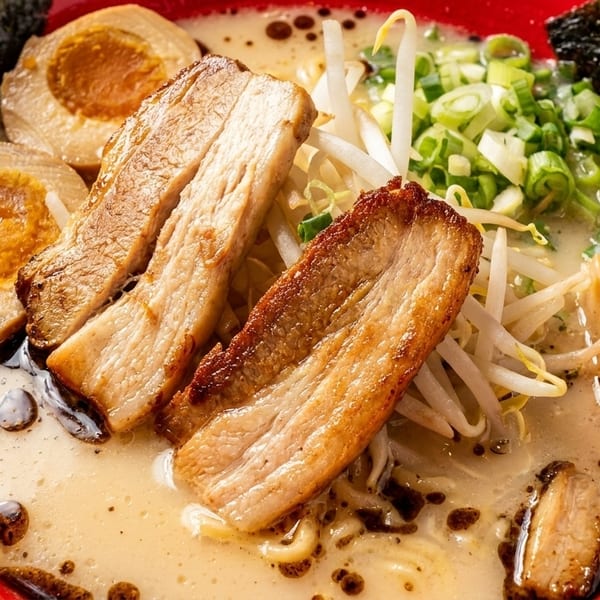 Close-up of ramen in a red bowl with pork belly, soft-boiled eggs, green onions, and nori.