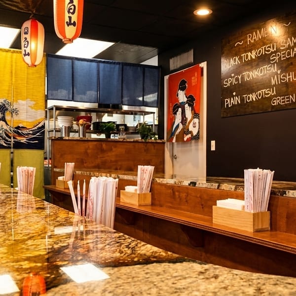 Interior of a Japanese restaurant with a long granite counter, menu board, and kitchen in the background.
