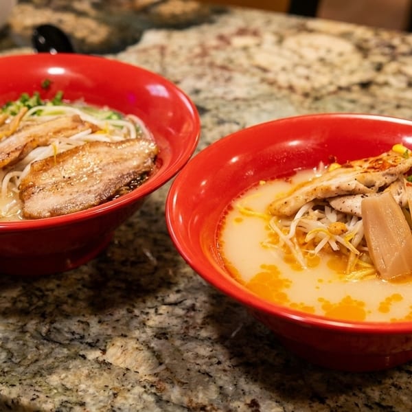 Two red bowls of ramen with various toppings, including pork, chicken, corn, and eggs.