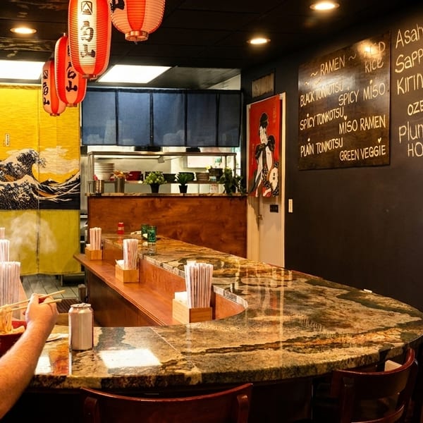 A man eating ramen at a curved counter in a Japanese restaurant with a chef in the background.