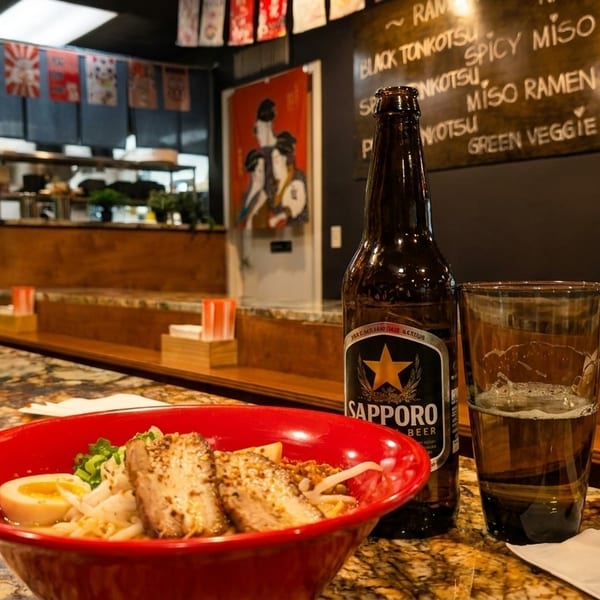 A red bowl of ramen with pork and egg, a Sapporo beer, and a glass of beer on a counter.